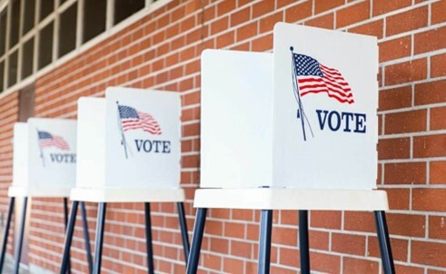 Voting booths along a brick wall. with an American flag and the words VOTE.